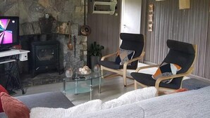 Un salón con chimenea de piedra, dos sillones negros, una mesa de centro de vidrio y un televisor sobre un escritorio.