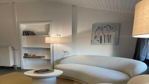 A modern living room with a beige sofa, a white coffee table, and a floor lamp. There is a bookshelf with books and decorative items, and a painting on the wall. - Sunny Cottage With Pool (Yvoir)