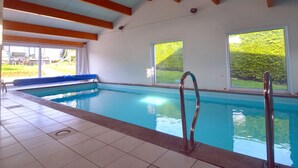 An indoor swimming pool with clear blue water, wooden beams on the ceiling, and large windows offering views of greenery outside. - Sunny Cottage With Pool (Yvoir)