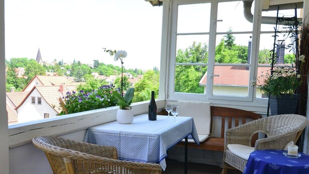 A balcony with wicker furniture, a table with a blue tablecloth, and a view of houses and greenery. - Apartment in Harz Near Ski Slopes (Quedlinburg)