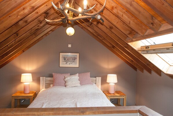 A cozy attic bedroom with a sloped ceiling, a chandelier, a bed with patterned pillows, and two bedside lamps.