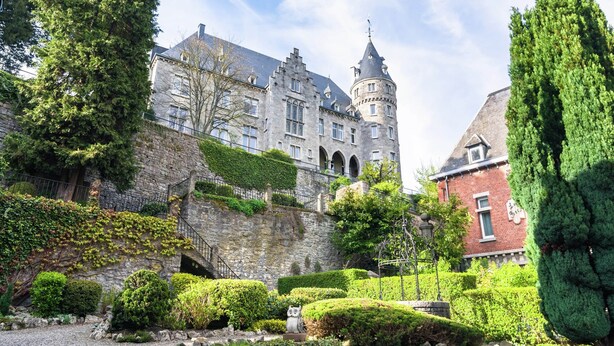 A historic building with a turret, stone walls, and a well-maintained garden with hedges and trees.