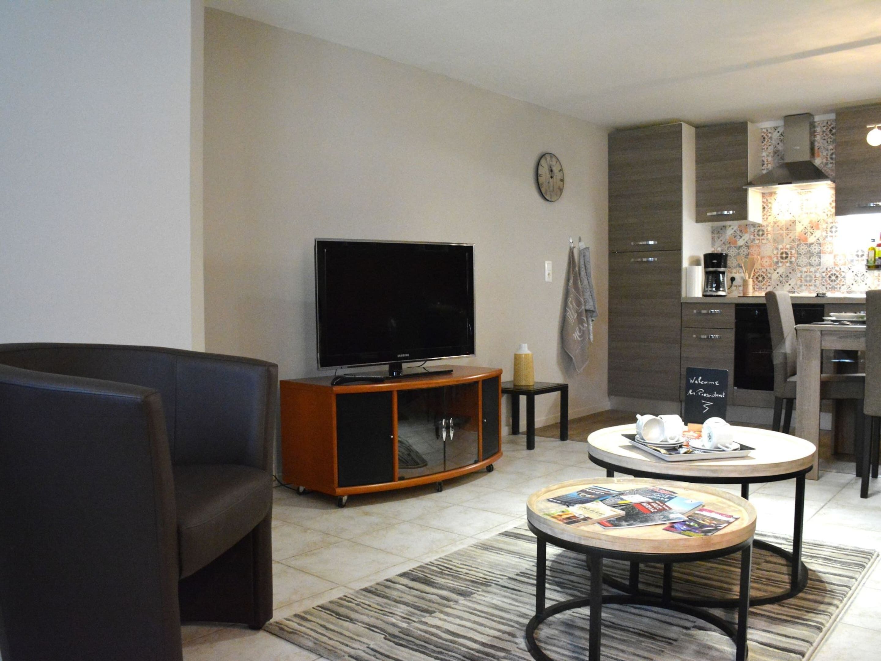 A modern living room with a flat-screen TV, a dark sofa, a coffee table with magazines, and a kitchen area with a tiled backsplash and a clock on the wall.