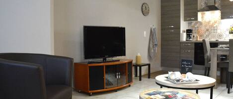A modern living room with a flat-screen TV, a dark sofa, a coffee table with magazines, and a kitchen area with a tiled backsplash and a clock on the wall.