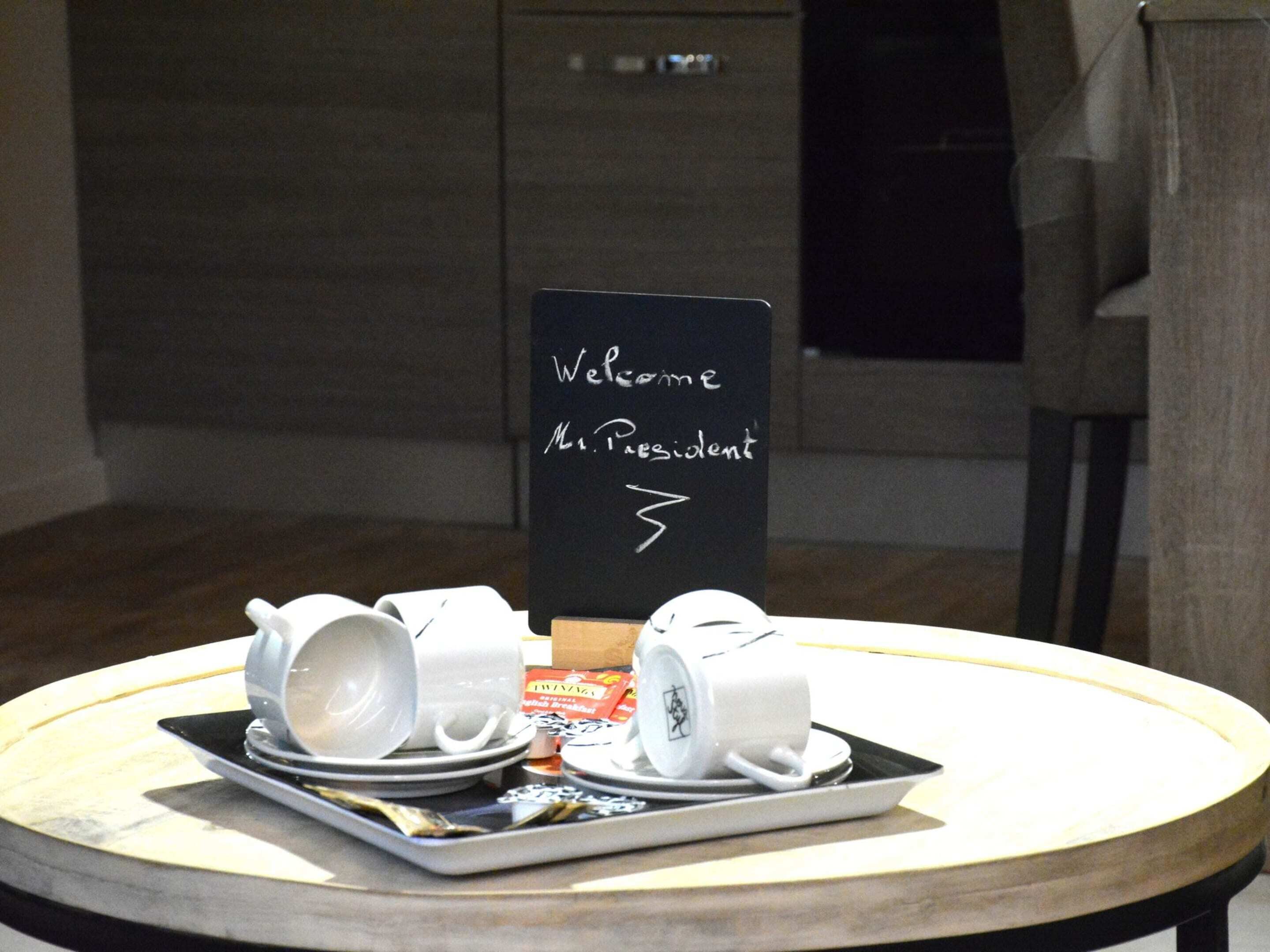 Une table ronde est dressée pour le petit-déjeuner avec un plateau contenant des tasses et des soucoupes blanches, ainsi qu’une petite boîte, qui contient probablement une boisson ou une pâtisserie. On peut voir, en arrière-plan, un panneau noir sur lequel est écrit : « Bienvenue, Monsieur le Président ».