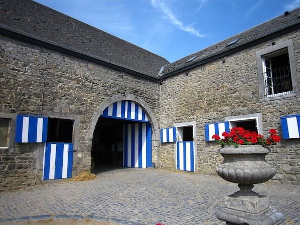A stone building with a cobblestone courtyard, a central arched doorway, and blue and white striped shutters on the windows.