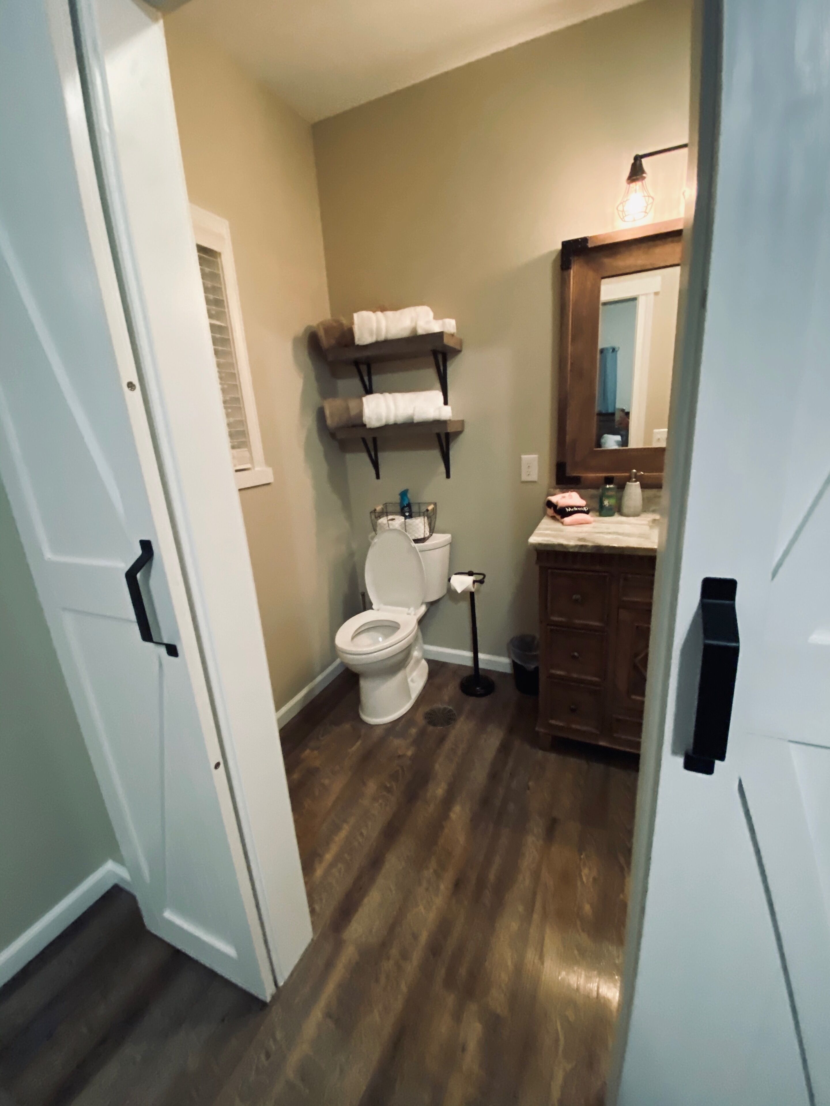 Une salle de bain avec des toilettes, un lavabo et un miroir. Des étagères se trouvent au-dessus des toilettes, où sont rangées des serviettes.