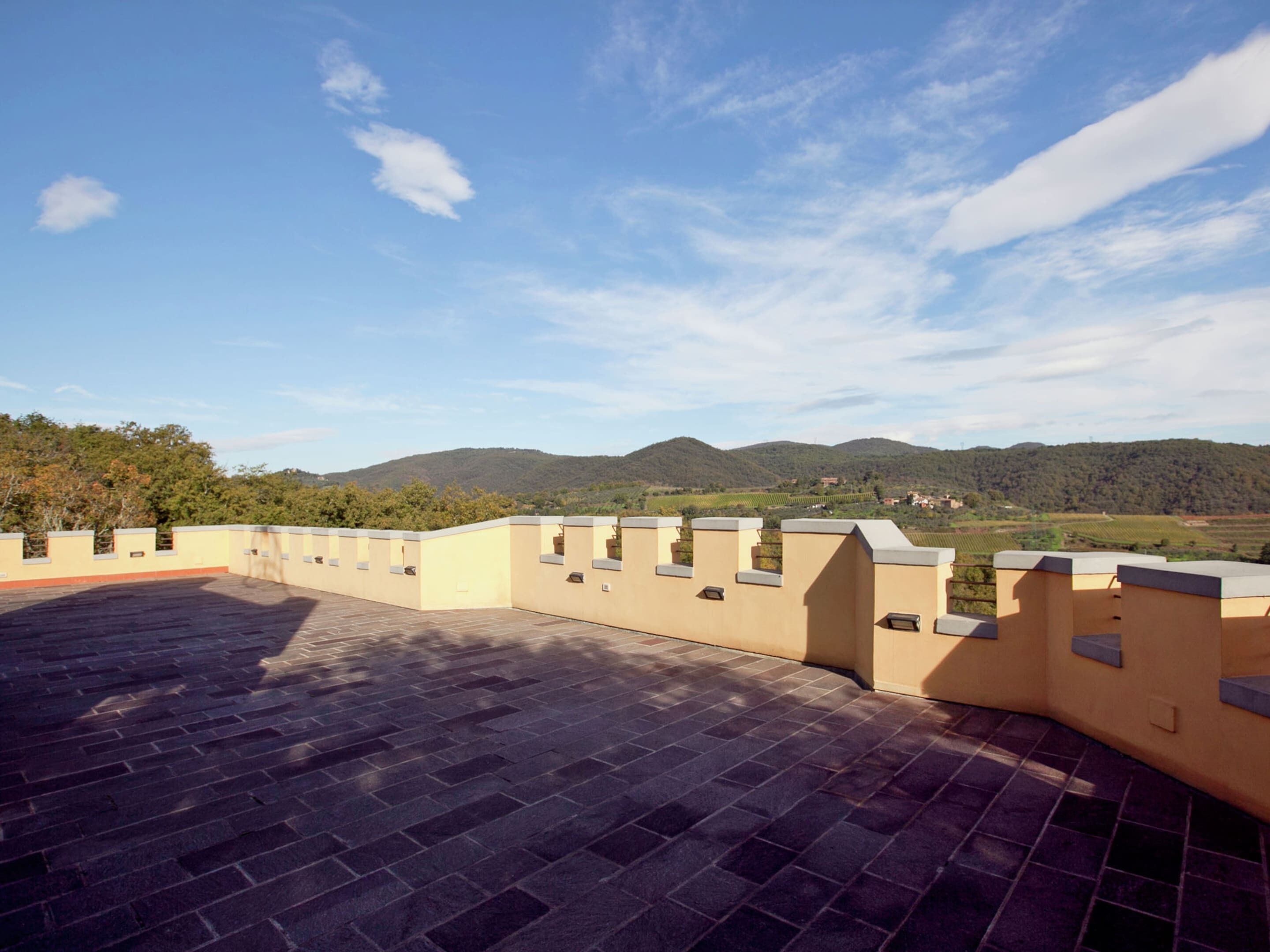 A rooftop terrace with a paved floor and a low wall, overlooking a hilly landscape.