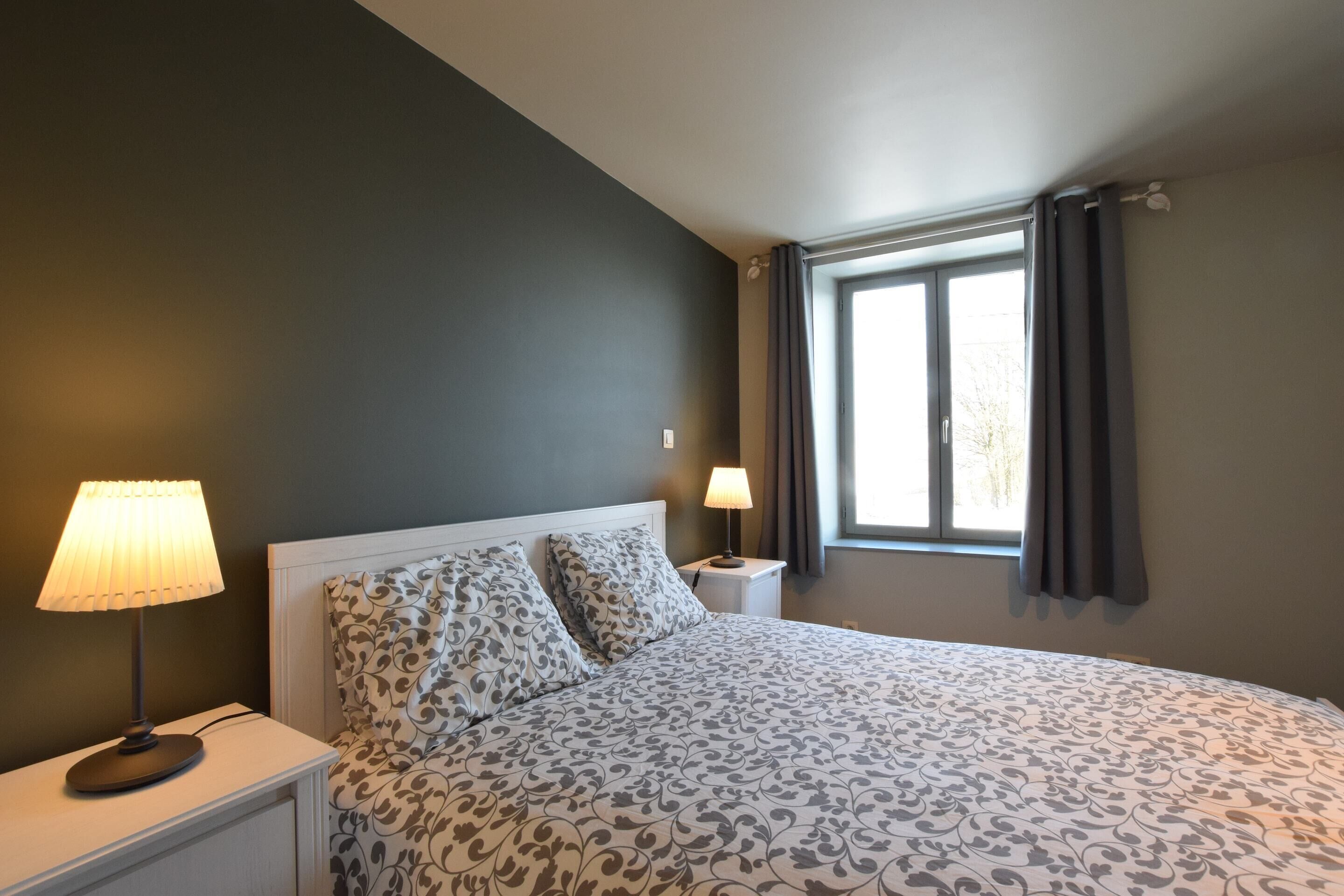 A bedroom with a bed, two bedside lamps, a window with curtains, and a nightstand.
