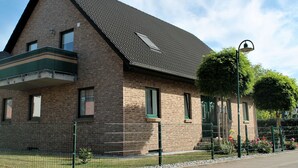 Ein Gebäude aus Backsteinen mit einem geneigten Dach, einem Balkon und einem von einer Hecke umgebenen Garten mit Blumen.