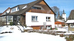 A two-story house with a wooden balcony, a visible chimney, and a church in the background. - Quiet Apartment in Merlsheim With Balcony (Nieheim)