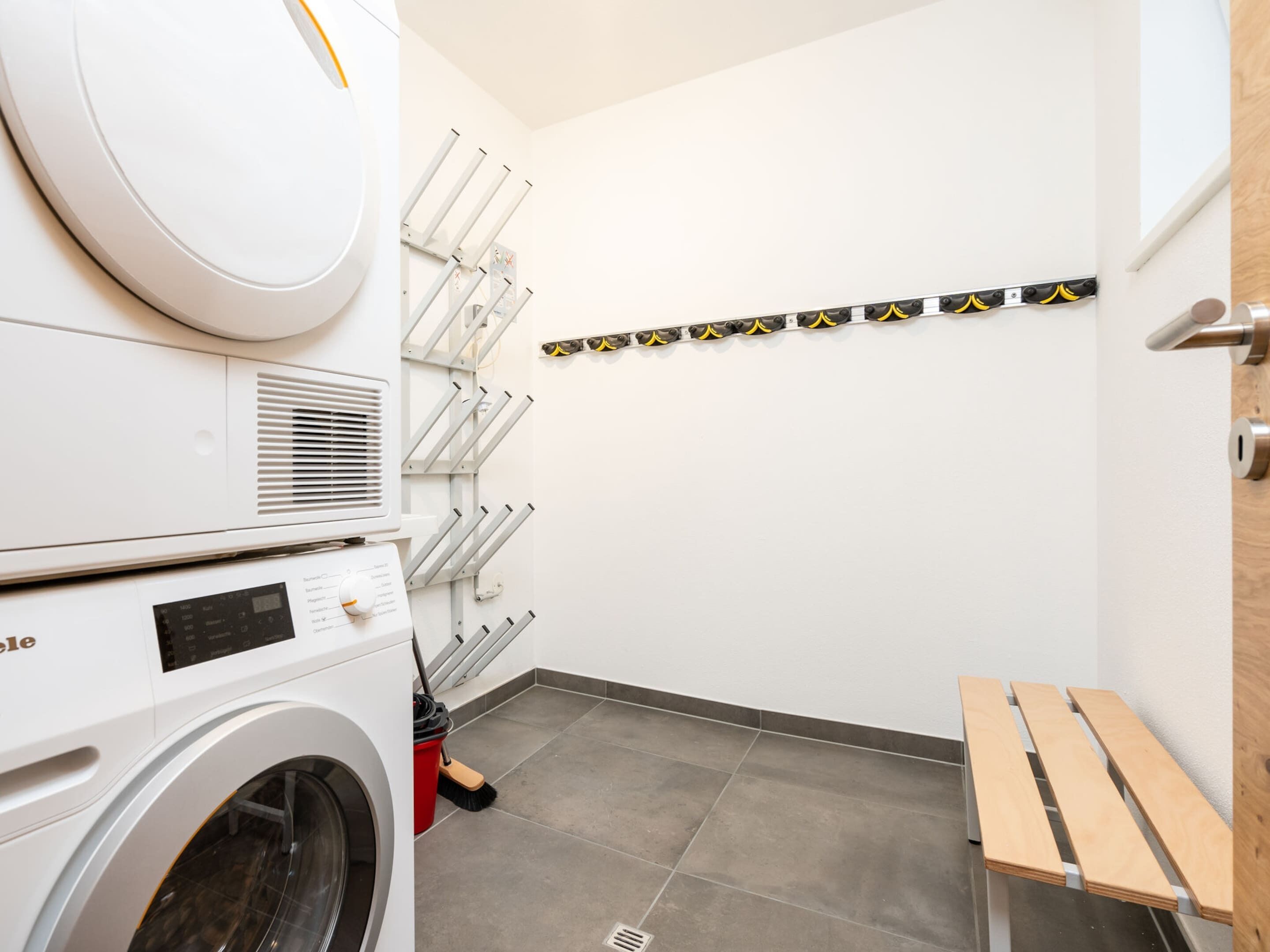 Une buanderie équipée d’un lave-linge à chargement frontal, d’un sèche-linge, d’un étendoir mural et d’un banc en bois.