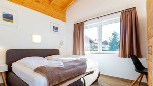 Ein Schlafzimmer mit Bett, Stuhl, einem Fenster mit Vorhängen und einer Holzbalkendecke.