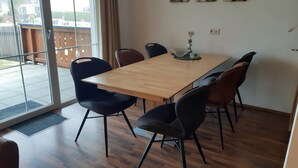Uma área de refeições com mesa de madeira e seis cadeiras, porta de vidro que dá para o exterior, e uma pequena mesa com um objeto decorativo.