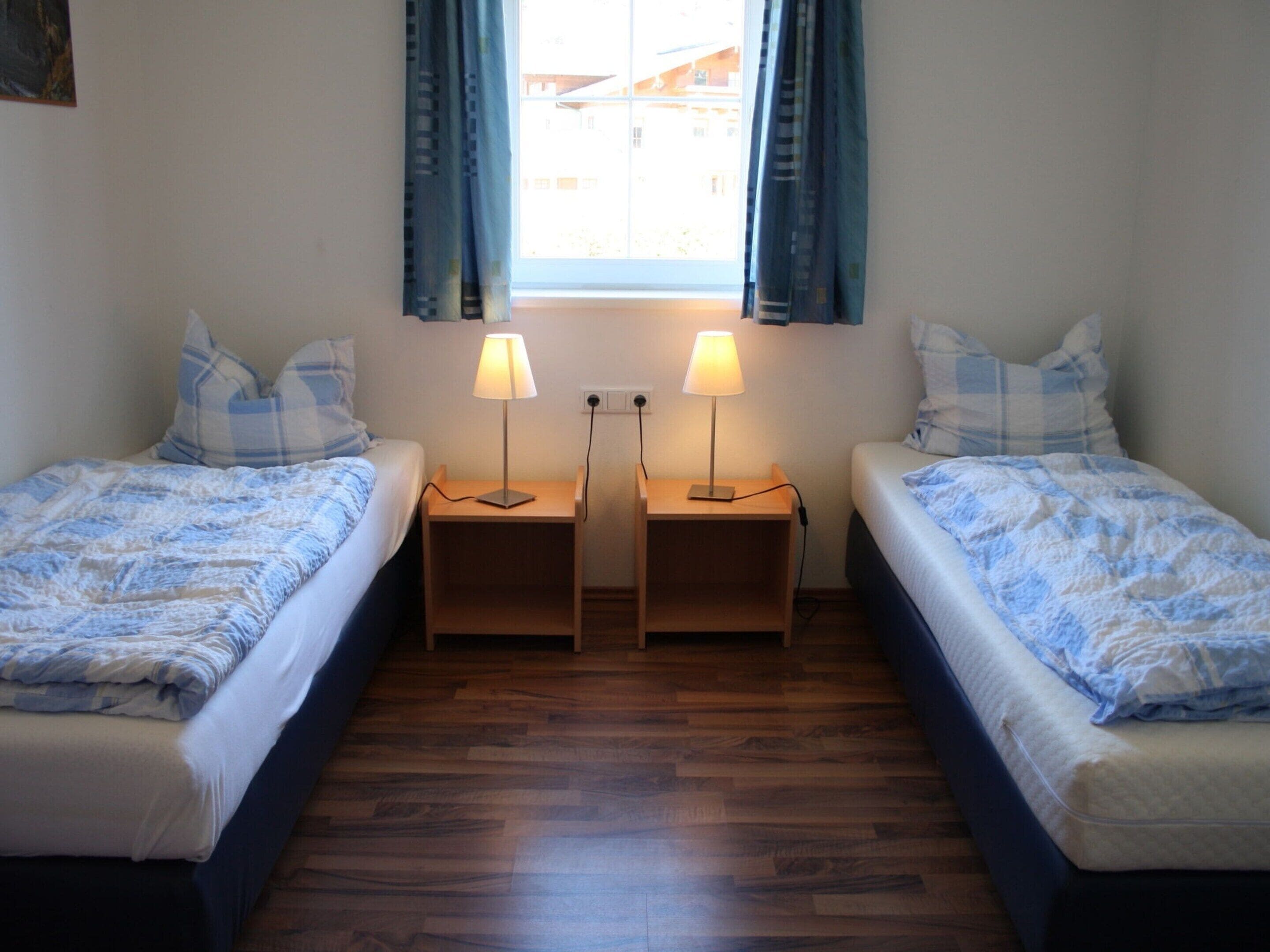 A room with two single beds, wooden nightstands, and a window with curtains.