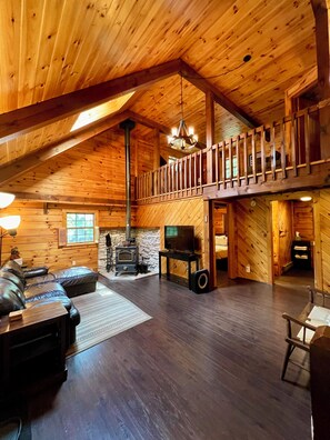 Un intérieur spacieux de chalet en bois, avec un plafond haut, une cheminée, un téléviseur et un coin salon confortable.