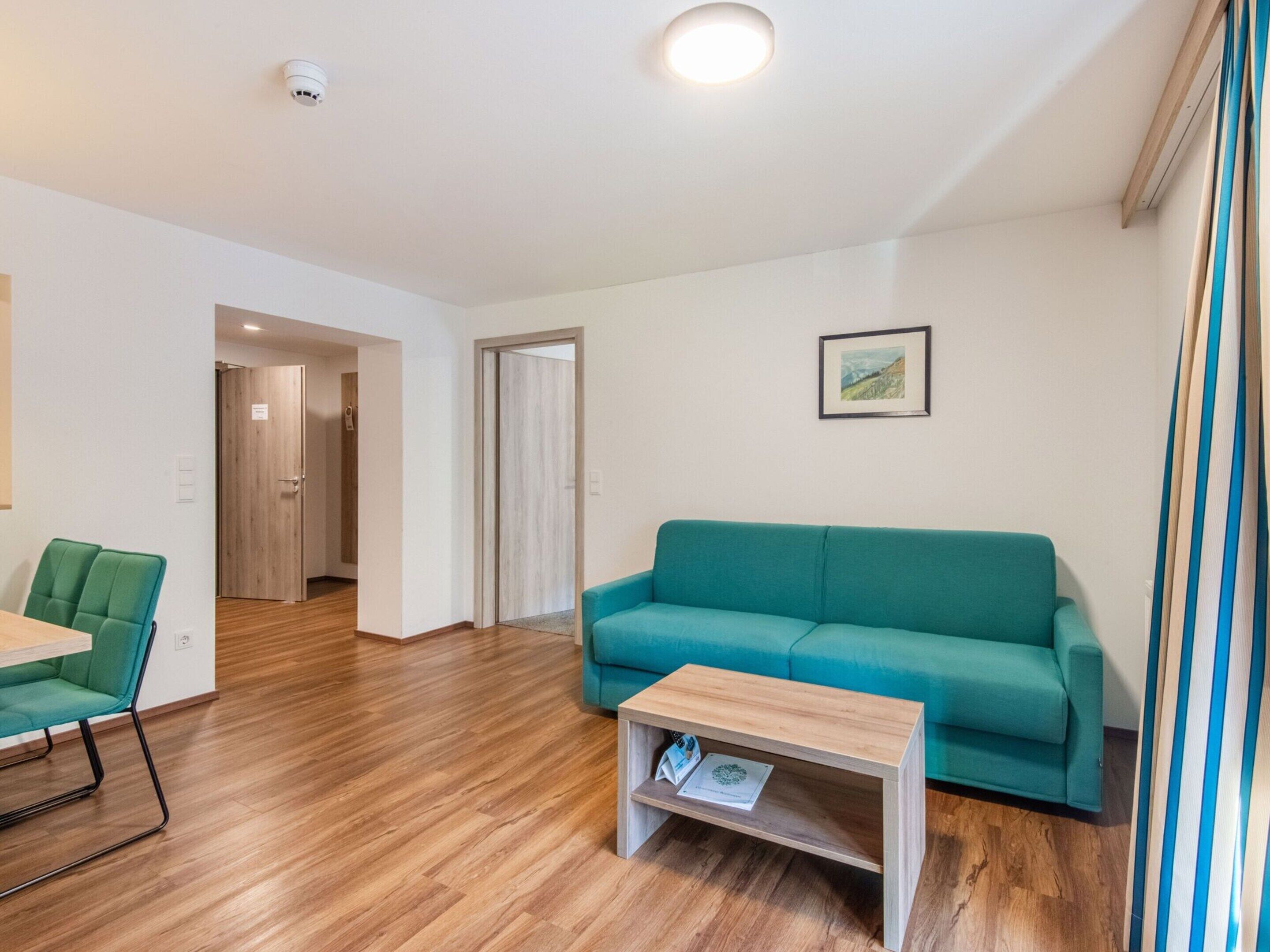 Un salon moderne avec un canapé de couleur turquoise, une table basse en bois et un fauteuil vert. Une porte donne accès à une autre pièce et un tableau encadré est accroché au mur.