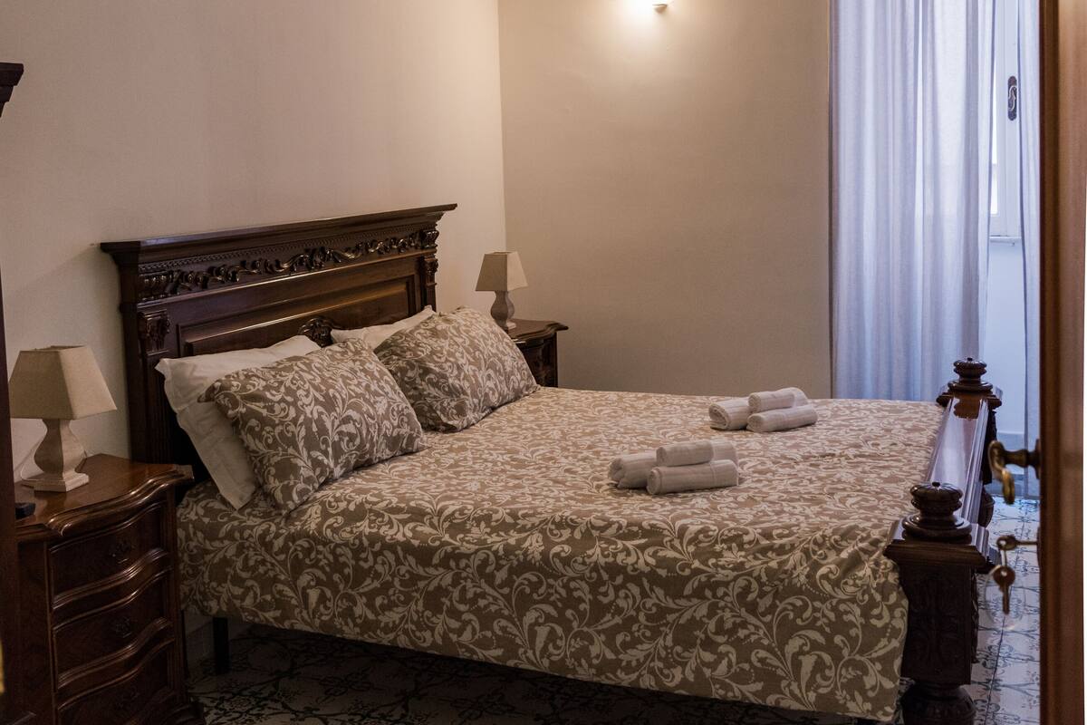 A bedroom with a bed, nightstands, a ceiling fan, a wall-mounted light, and a window with curtains.
