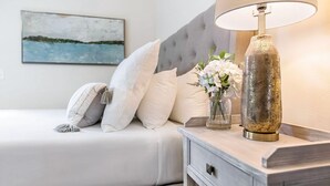 Una cama bien hecha con ropa de cama blanca, un cabecero gris con capitoné y una mesita de noche con una lámpara y un florero.