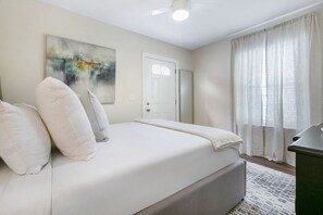 Un dormitorio con cama, una puerta, una ventana con cortinas y un cuadro en la pared.