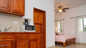A kitchen with wooden cabinets, a microwave, and a coffee maker. There is a bed with a pink blanket in a room with a ceiling fan and a window with curtains. - The Orchid Studio Experience" (Caye Caulker)