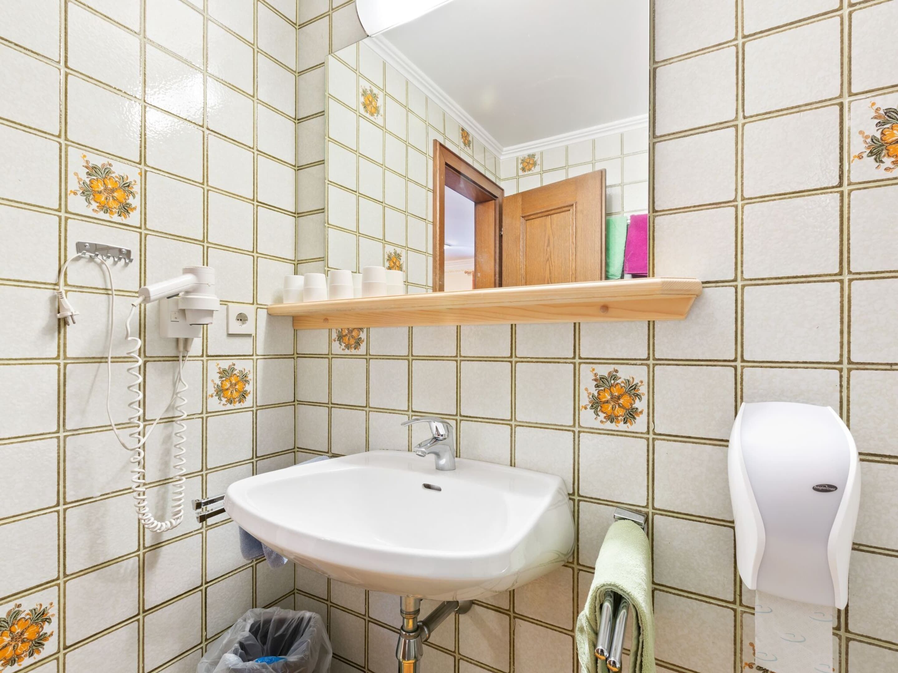 A bathroom with a white sink, a mirror, a towel dispenser, and a shelf with toiletries.