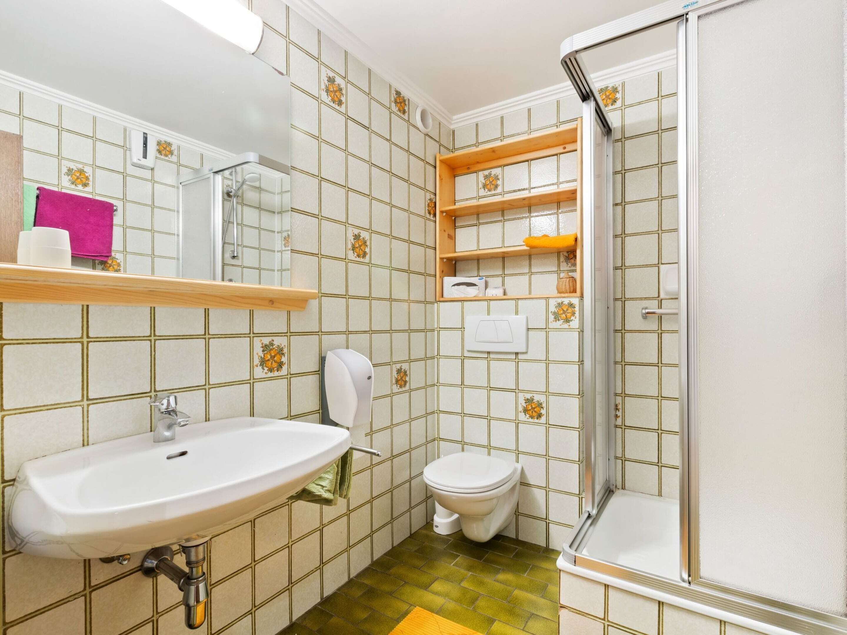 Een badkamer met een wastafel, toilet en douche. Er zijn planken boven het toilet en een spiegel boven de wastafel.