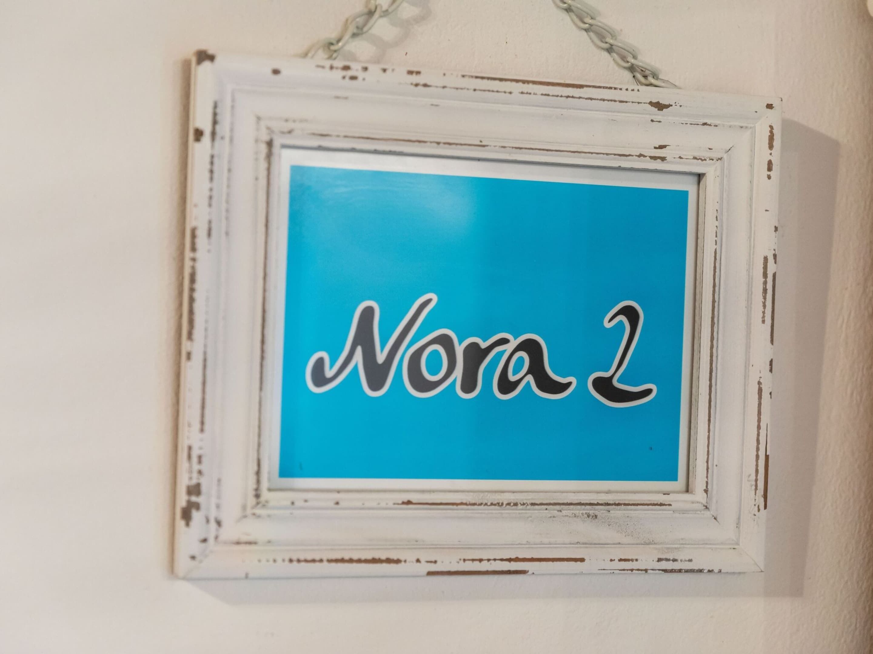 Een lijst met een bord waarop de tekst 'Nora 2' staat, tegen een blauwe achtergrond.