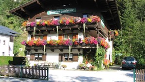 Un chalet traditionnel doté de plusieurs balcons ornés de fleurs colorées, une enseigne indiquant qu’il s’agit d’une pension, et une voiture garée devant.