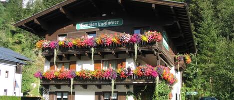 A traditional chalet with multiple balconies adorned with colorful flowers, a sign indicating it is a pension, and a car parked in front.