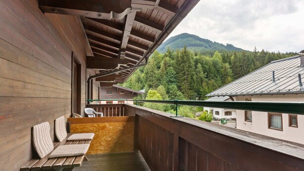 Un balcon en bois avec un banc, une vue sur une montagne boisée et un bâtiment doté d’un toit en pente.