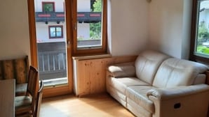A room with a wooden floor, a beige sofa, a wooden chair, and a window with a view of a building with balconies. - Spacious Apartment in Salzburg near Ski Area (Saalbach-Hinterglemm)