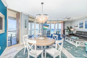 Ein geräumiges Wohnzimmer mit einem runden Holztisch, weißen Stühlen, einer großen Chandelüster, einem Sofa und Meerblick durch eine Schiebeglastür.