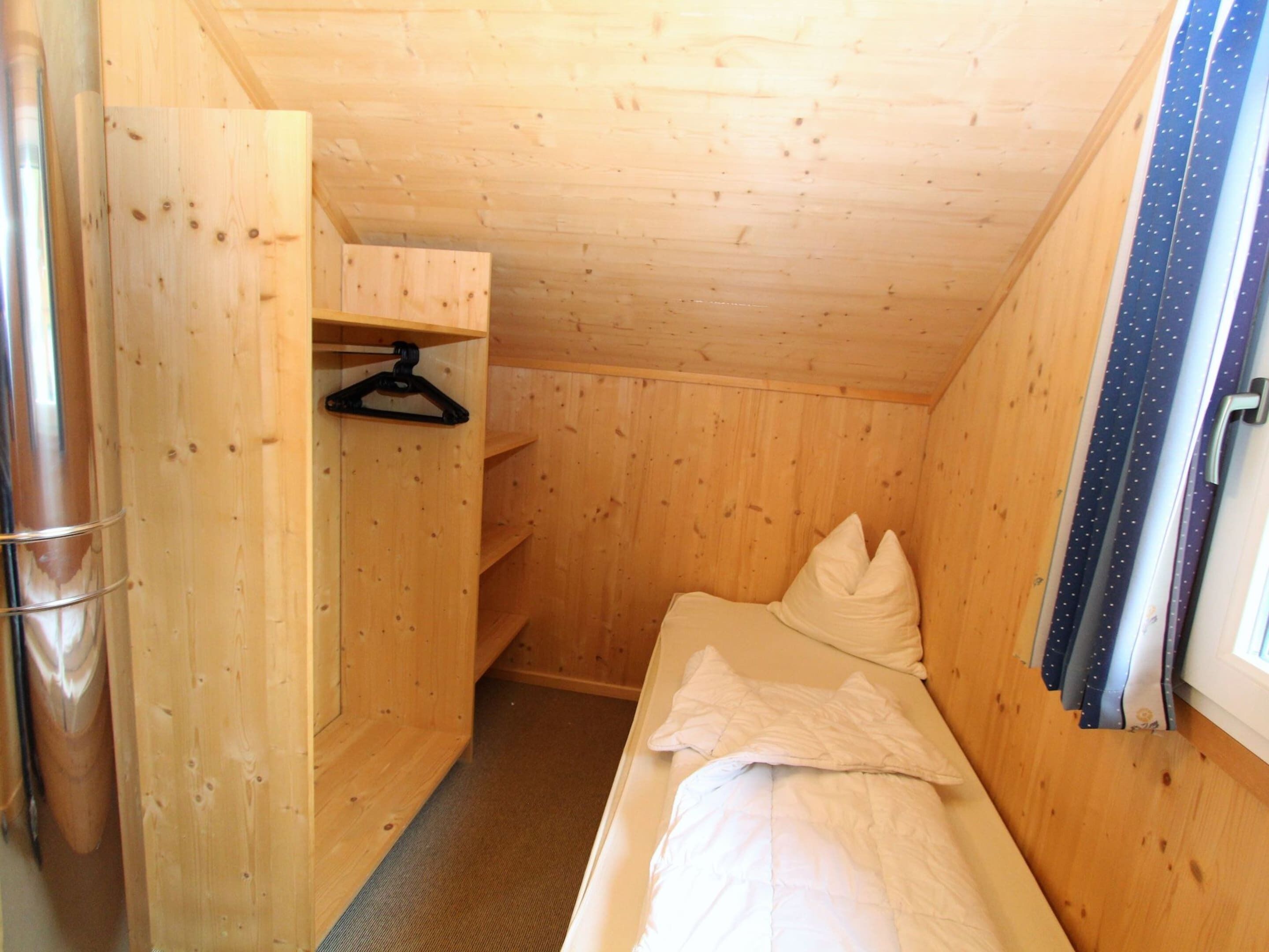 A small, wooden cabin room with a single bed, a hanging clothes rack, and a small shelf.