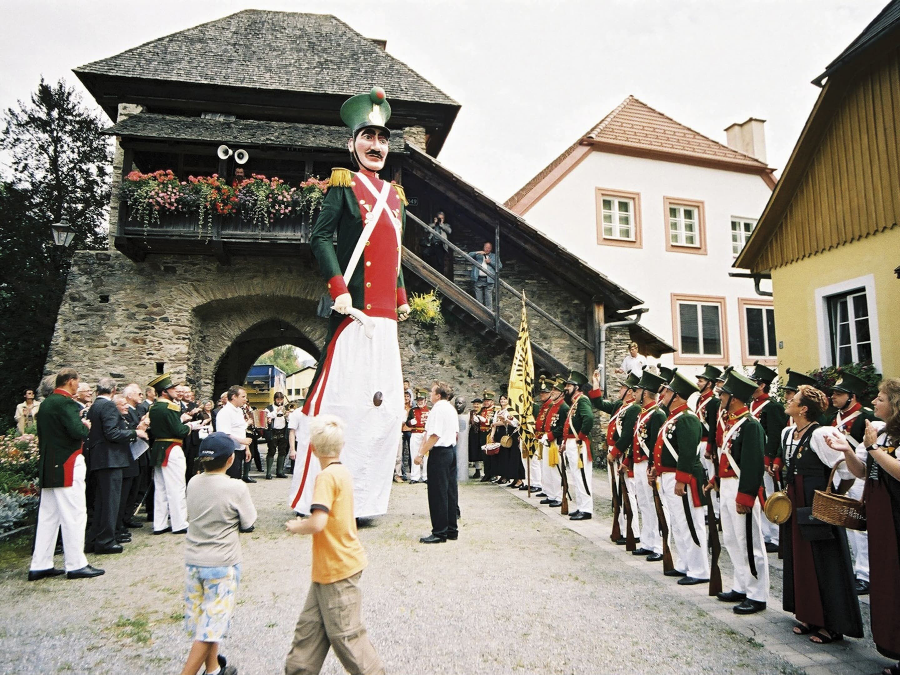 A historical reenactment with participants in period costumes, a large figure in a uniform, and a crowd observing.