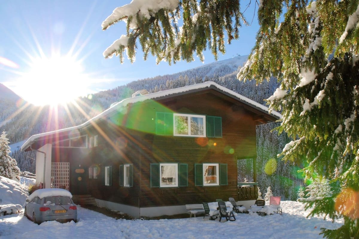 A wooden cabin with a sloped roof, surrounded by snow-covered ground and trees, with a clear sky and sunlight.