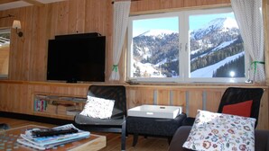 Un salon chaleureux avec des murs en bois, un téléviseur à écran plat, une bibliothèque et une fenêtre donnant sur un paysage de montagne enneigé.