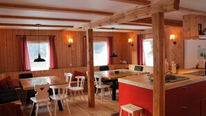 L’intérieur chaleureux d’un chalet avec des murs en bois, un coin repas avec des chaises blanches, une cuisine dotée d’un plan de travail rouge et un petit tabouret.