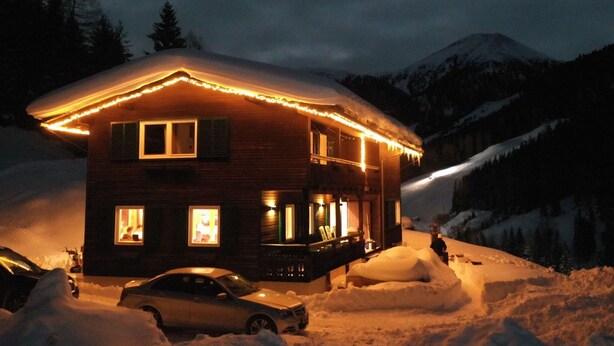 Une cabane en bois, dont le toit est recouvert de neige et dont l’extérieur est illuminé, entourée d’un paysage enneigé et de montagnes.