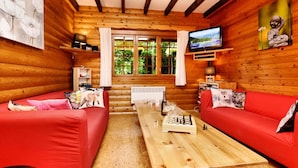 L’intérieur d’un chalet accueillant, avec des murs et un plafond en bois, un téléviseur fixé au mur, une cheminée et une grande fenêtre agrémentée de rideaux.
