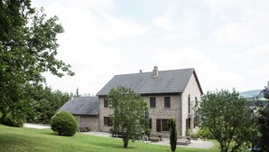 Une maison en pierre à deux étages, dotée d’un toit en pente, entourée d’une pelouse bien entretenue et de plusieurs arbres.