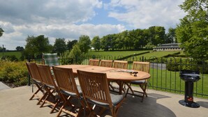 Een terras buiten met een ronde houten tafel en zes houten stoelen met kussens.