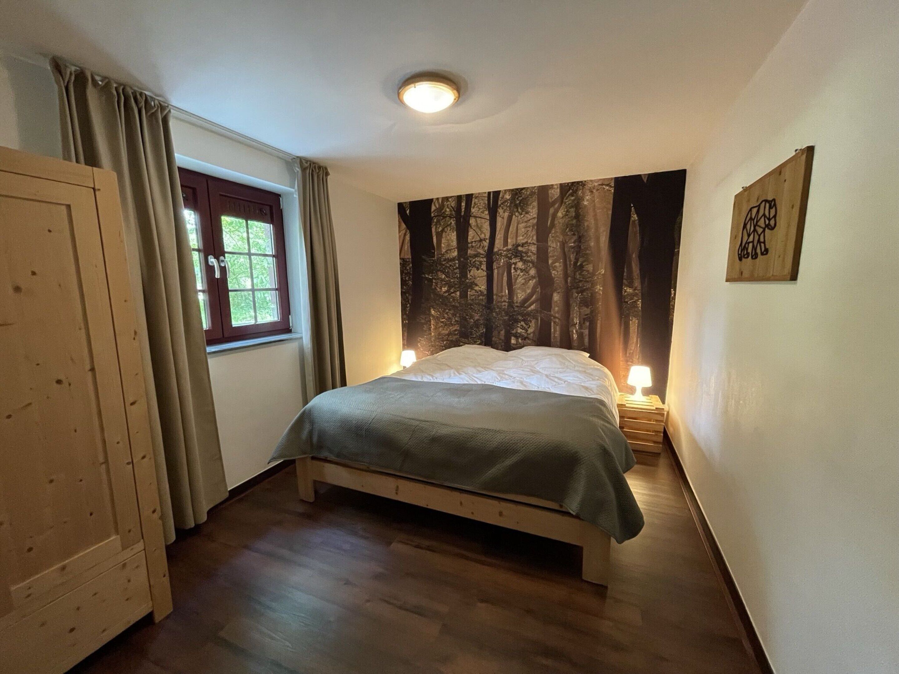 Um quarto com uma cama, uma janela com cortinas, uma parede com um mural de floresta e uma porta de madeira.