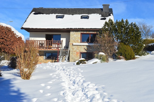 A two-story house with a stone facade, a wooden deck, and a snow-covered driveway.