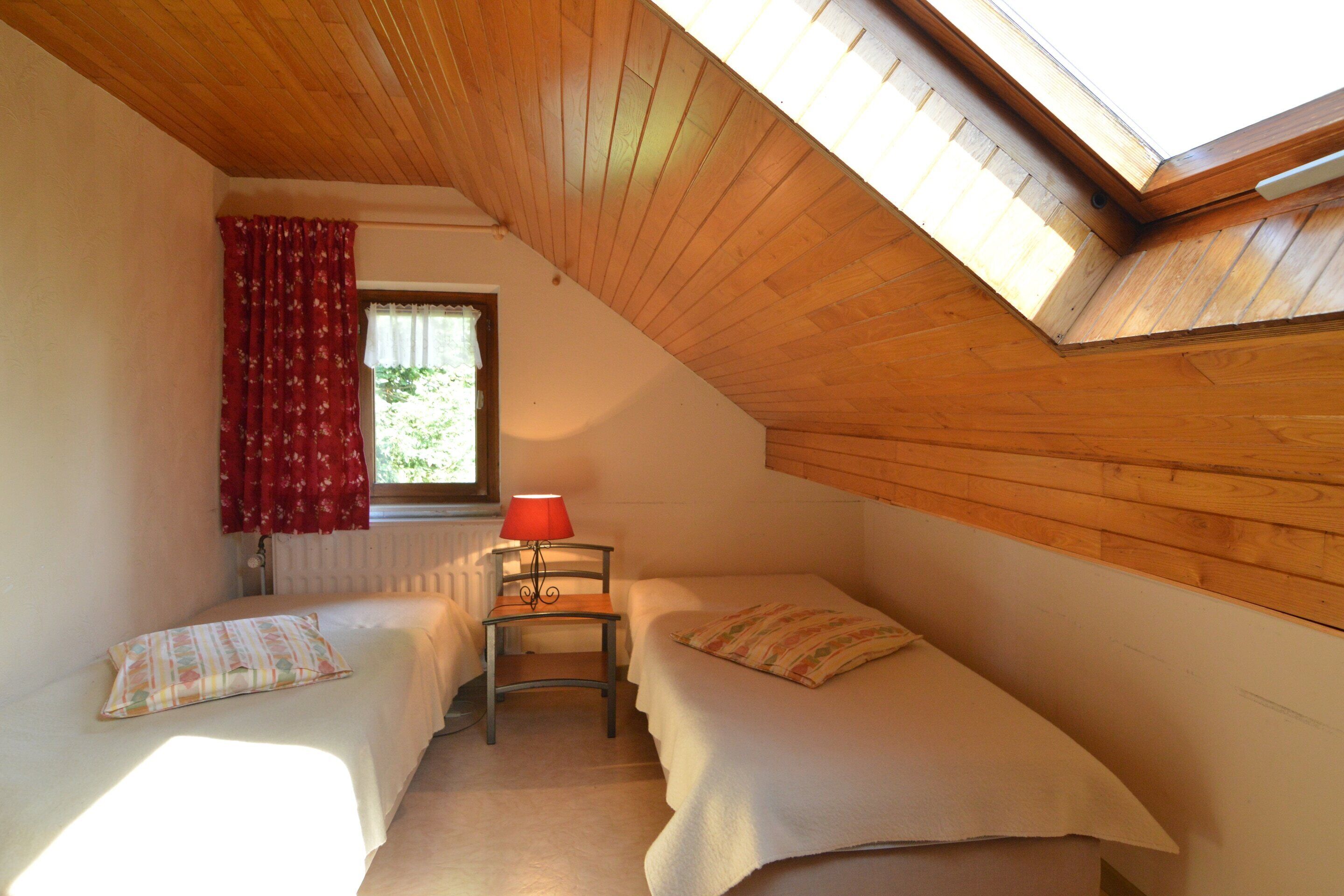 A room with two beds, a window with red curtains, a table with a lamp, and a sloped wooden ceiling.