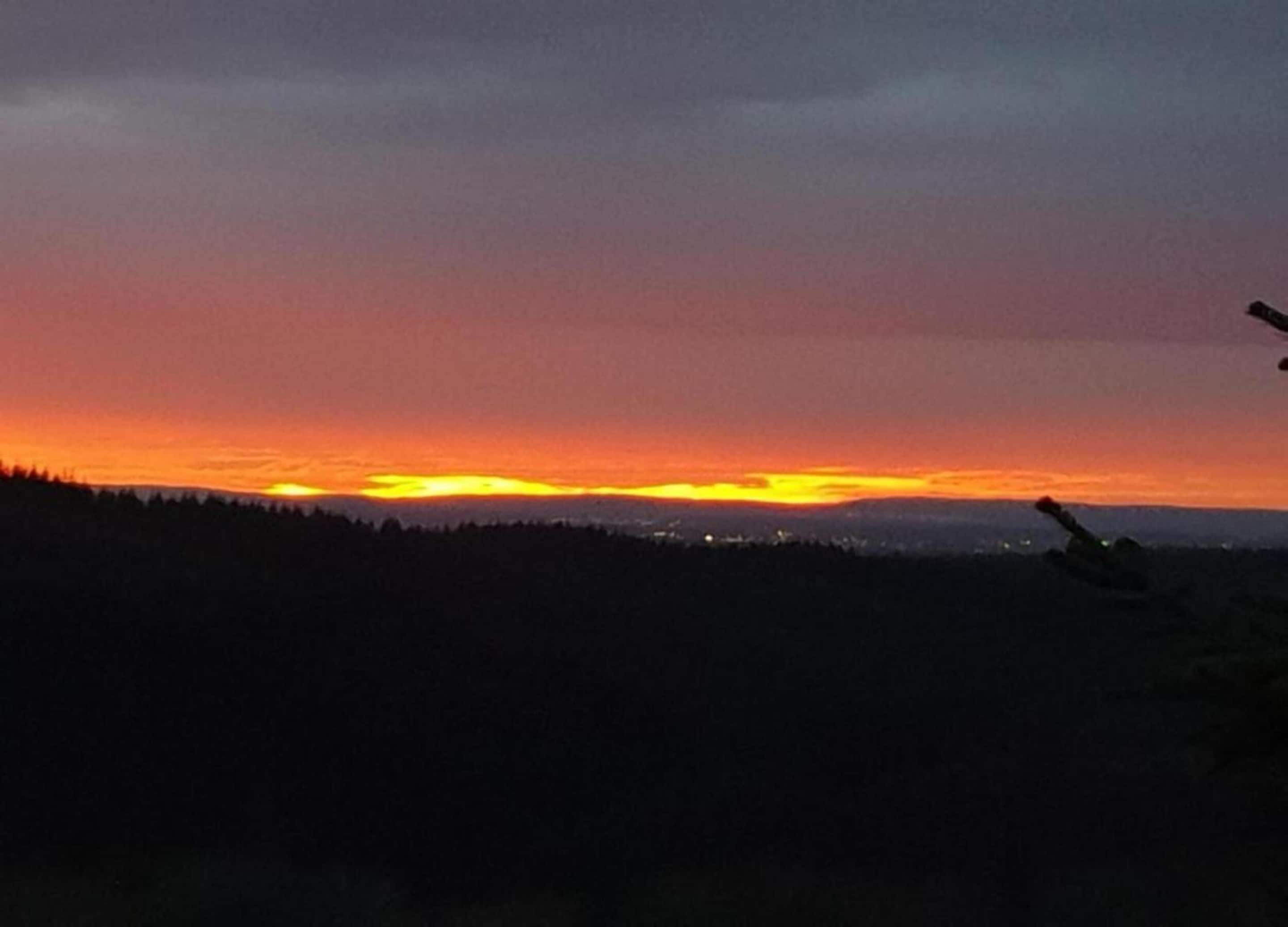 A sunset over a forested landscape with a silhouette of a person in the foreground.