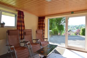 Ein Zimmer mit vier braunen Ledersesseln, einem kleinen Tisch mit einer verzierten Fliese, einem Fenster mit Vorhängen und Blick auf einen Parkplatz und Grünflächen.
