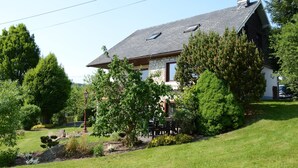 A house with a sloped roof, stone foundation, and a well-maintained garden with various trees and shrubs. - Ondenval Stay With Sauna (Waimes)