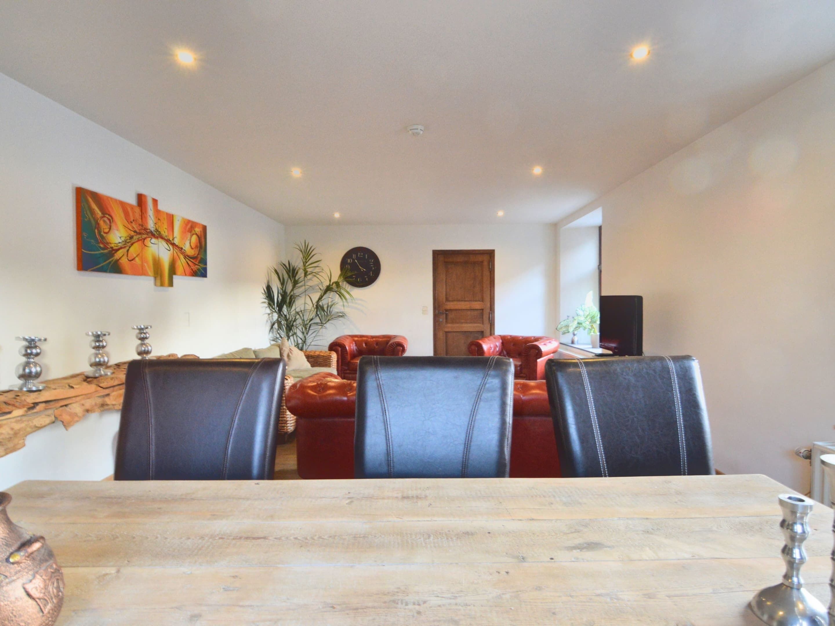 A dining area with a wooden table and four chairs, a red sofa, a clock on the wall, and a plant.