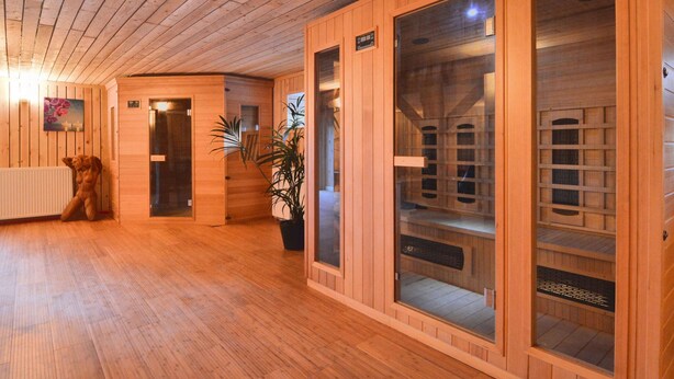 Ein Saunabereich mit Holzwänden und Holzboden. Zwei Saunakabinen mit Glastüren und Sitzbänken sind vorhanden. Vor dem Eingang steht ein Blumentopf.